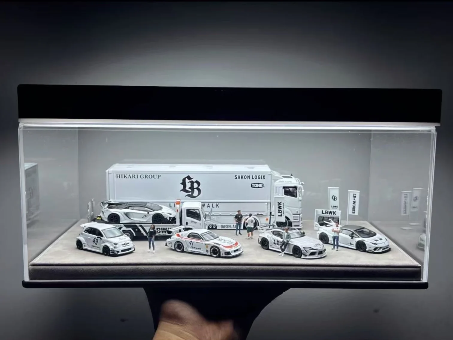 A curated 1:64 scale JDM model car display organized by theme, featuring white Liberty Walk (LBWK) cars and a transport truck in an acrylic case.